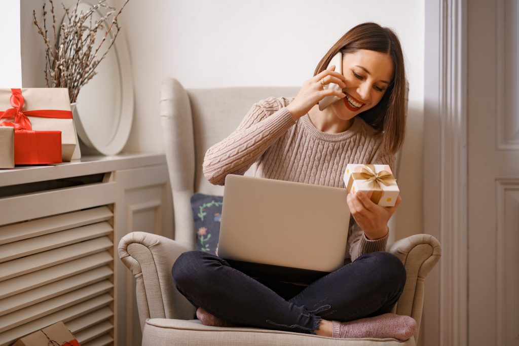 Laughing young woman with laptop talking on mobile phone while sitting in a comfortable armchair holding a gift box surrounded by gifts at home